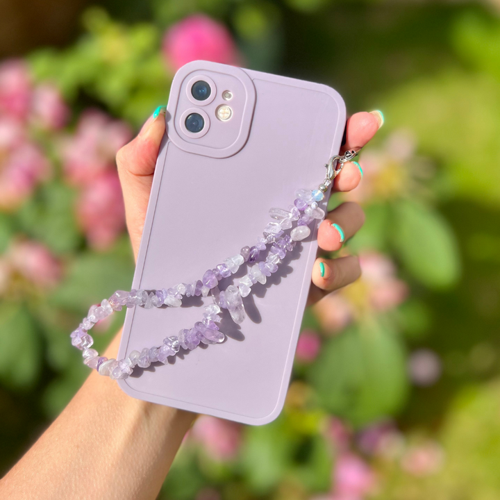 A hand holding a smartphone encased in the NAKD Phone Case - Lilac from Coconut Lane’s spring collection. Attached to the case is a beaded wrist strap crafted from purple and white stones. The blurred greenery and pink flowers in the background beautifully capture the essence of pastel favourites.