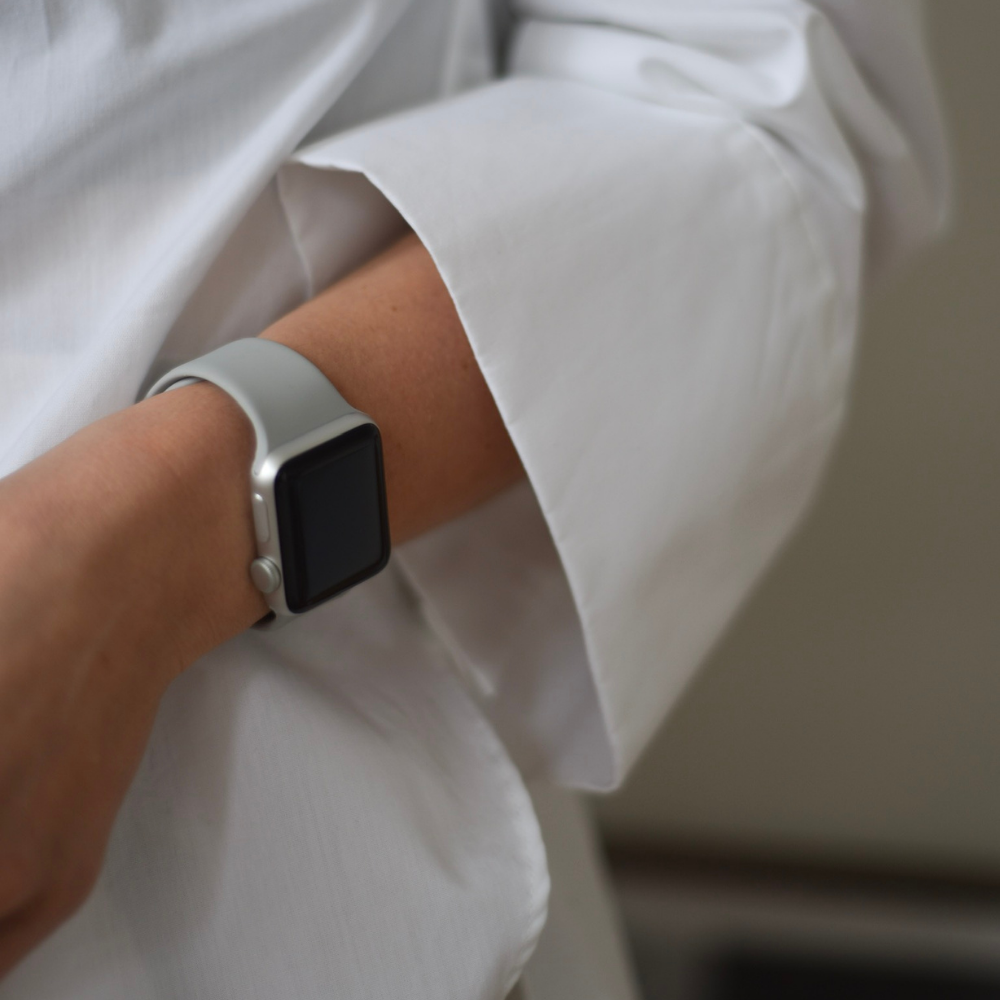 Close-up of a person wearing a white long-sleeve shirt and a Coconut Lane NAKD Apple Watch Strap in Ice Grey on their wrist. The person’s arm is slightly bent, and the smartwatch screen is off. The image focuses on the wrist area, highlighting the stylish strap from Coconut Lane's NAKD Collection.