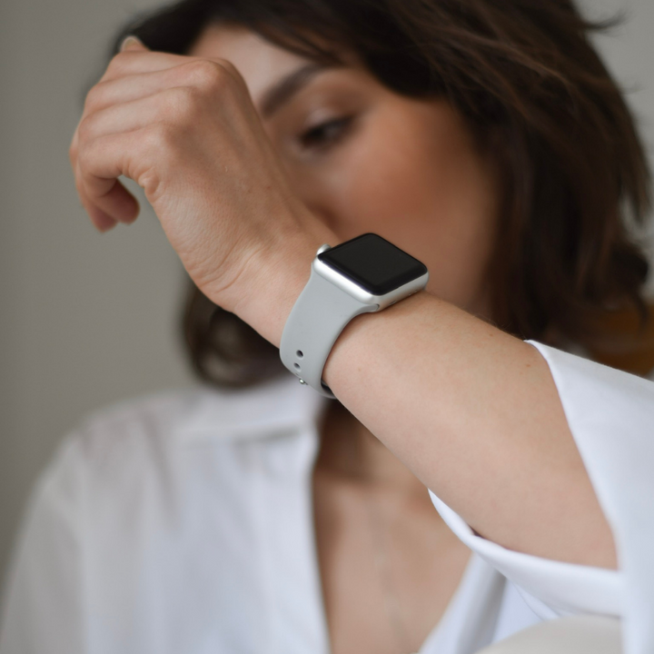A person with short brown hair, wearing a white shirt, is holding their wrist up to their face, showcasing a NAKD Apple Watch Strap in Ice Grey by Coconut Lane. The smartwatch face is not visible as the background is blurred, emphasizing the wrist and watch strap.