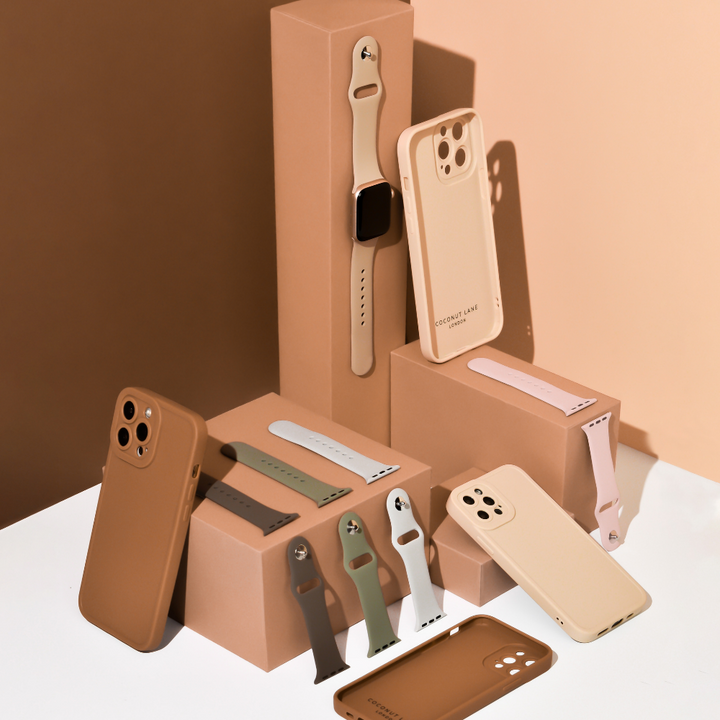 A collection of phone cases and watch bands in various shades of beige, tan, green, and pink are displayed on stacked brown boxes. An assembled beige smartwatch with a NAKD Apple Watch Strap - Ice Grey from Coconut Lane is also visible, standing upright on one of the boxes. The background features neutral tones.
