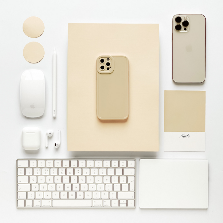 A neatly arranged flat lay composition features a beige iPhone with a Coconut Lane NAKD Phone Case - Nude offering full camera protection, another beige iPhone, an Apple Magic Mouse, a white keyboard, AirPods and individual pieces, a stylus, a beige notepad, two coasters, and a color swatch labeled "Nude.