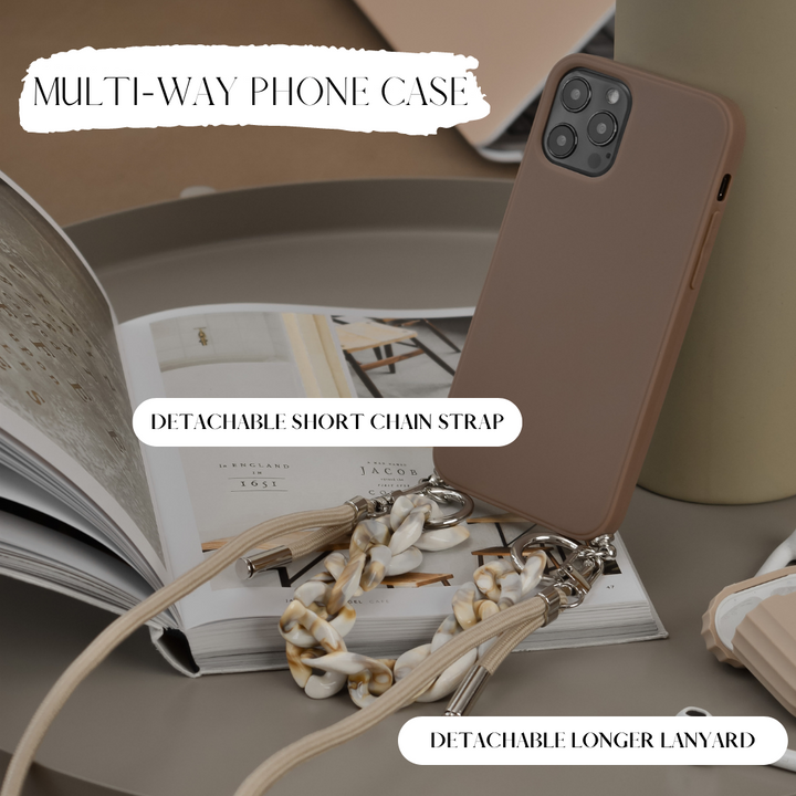A smartphone in the Lanyard & Chain Phone Case - Brown by Coconut Lane, featuring a multicolored chain strap and a beige cord lanyard, lies on an open book on a table. The image highlights the detachable short chain strap and detachable longer lanyard. Text reads "MULTI-WAY PHONE CASE.