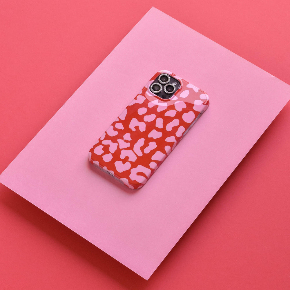 A smartphone with a red and light pink abstract patterned case, featuring tempered glass and Coconut Lane's Protective Camera Lens Covers - Diamante for the iPhone, is placed on a slightly tilted pink surface, against a coral red background. The colors of the scratch-resistant phone case complement the shades of the pink and coral red backdrop.