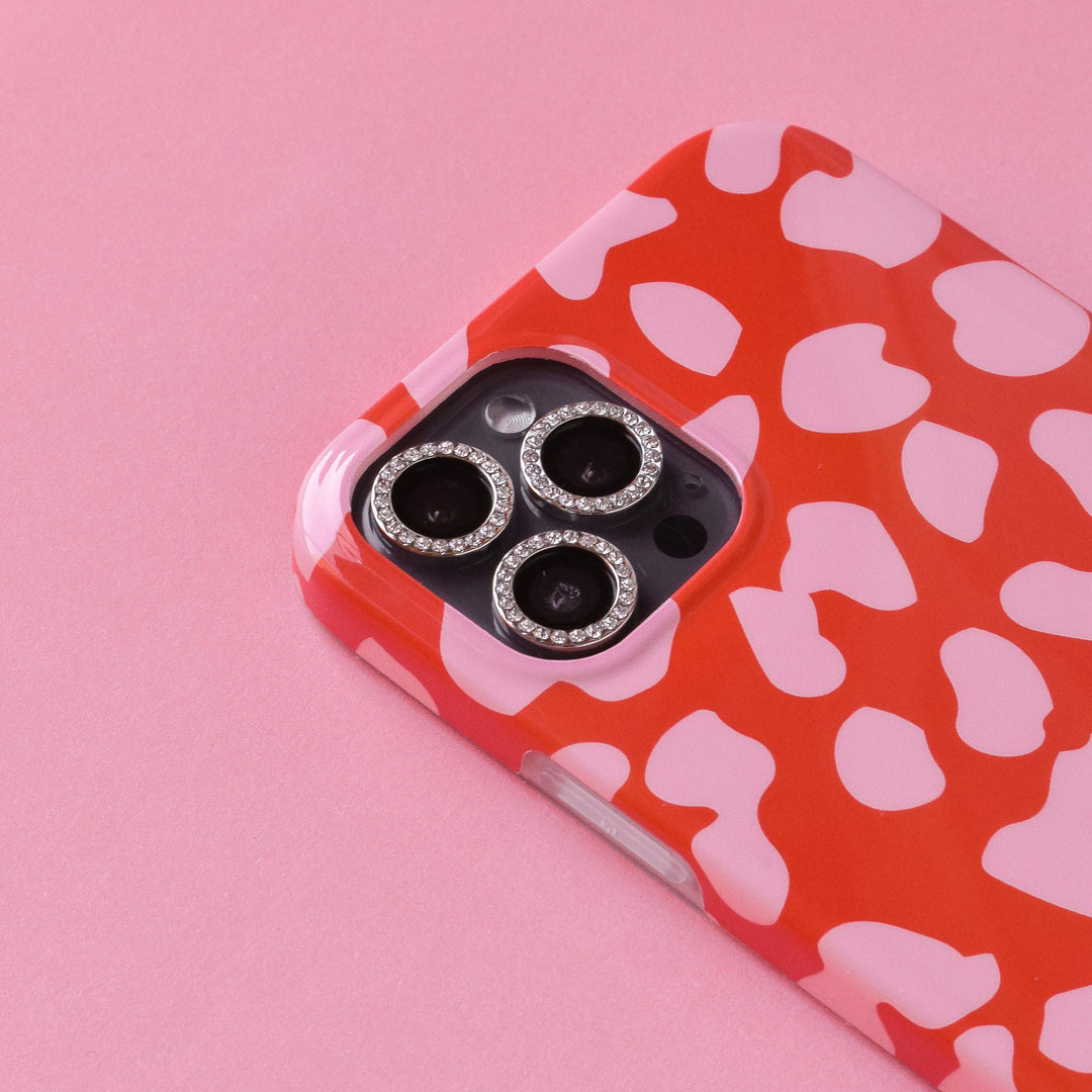 A close-up of a smartphone with a red and white leopard print case on a pink background. The Coconut Lane Protective Camera Lens Covers - Diamante feature diamond-like embellishments, adding a touch of sparkle to the device while ensuring it remains scratch-resistant.