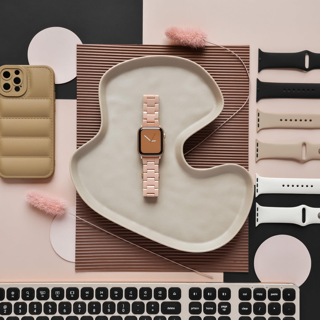 A flat lay of a pink smartwatch with a matching band on a wavy tray, surrounded by various colored smartwatch bands including the Luxe Nude Apple Watch Strap from Coconut Lane, a smartphone with a tan case, a keyboard, and pink fluffy decorative elements, all set against a beige and brown geometric background.