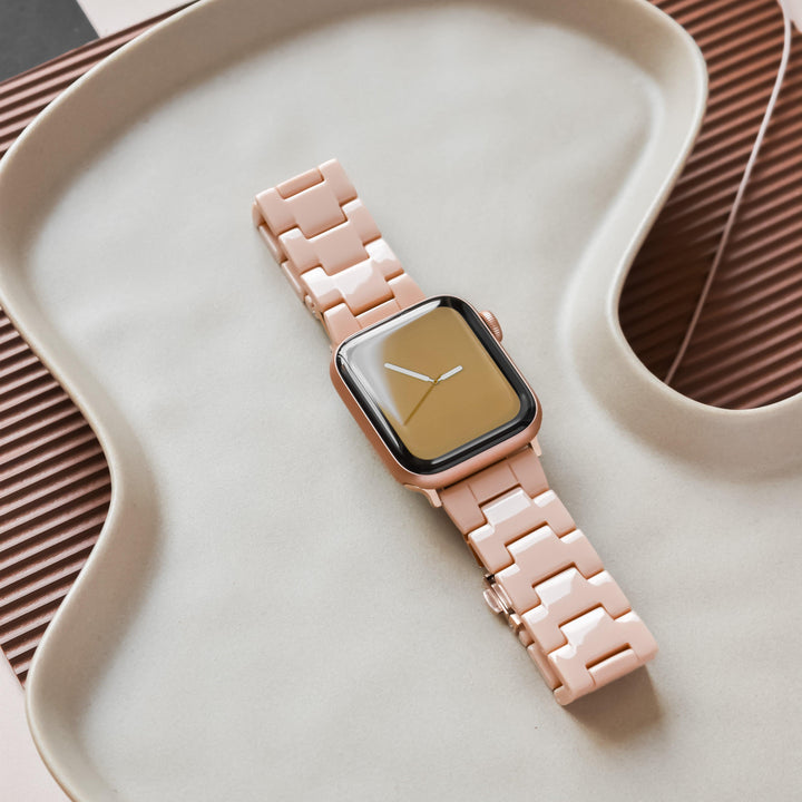 A Luxe Nude Apple Watch Strap by Coconut Lane, featuring a pink, segmented design, is elegantly displayed on a curved beige dish. The watch face is dark and reflective, displaying the time with minimalist white hour and minute hands. The backdrop showcases alternating textured surfaces in brown and beige tones.