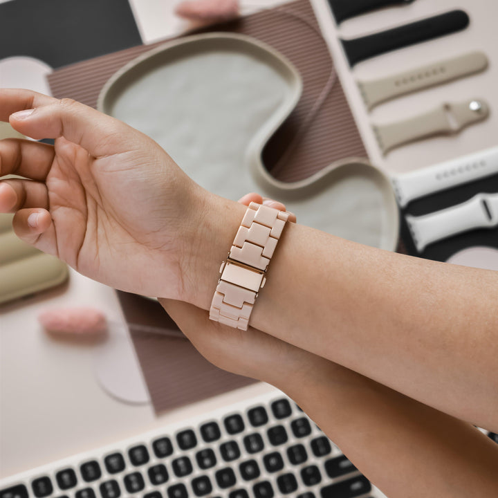 A close-up of a hand wearing the Luxe Nude Apple Watch Strap from Coconut Lane, featuring an adjustable design in beige. The background includes a laptop keyboard, various Luxe Nude Apple Watch Straps in different colors, a sleep mask, and other personal items arranged on a desk.