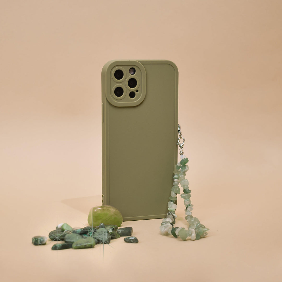 A Crystal Phone Strap - Green by Coconut Lane is displayed on a beige background, showcasing its beaded charms attached to a green phone case. Small green stones and a polished Green Aventurine gemstone are scattered beside it, creating a minimalist and earthy aesthetic.
