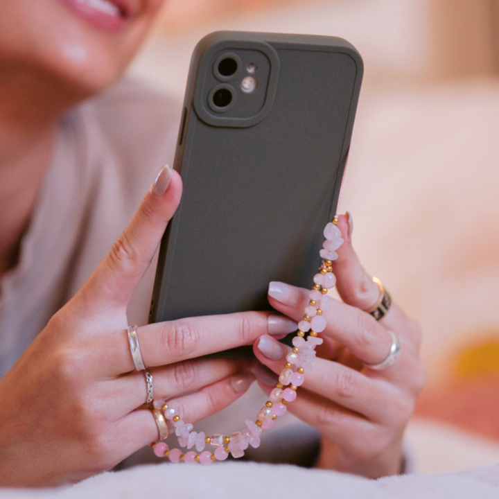 A person is holding a black smartphone with a pink and gold beaded phone strap attached to their Coconut Lane NAKD Phone Case in Khaki. The individual has manicured nails with light pink nail polish and is wearing multiple rings on their fingers. The background is blurred, focusing on the hands and phone, emphasizing minimalism.