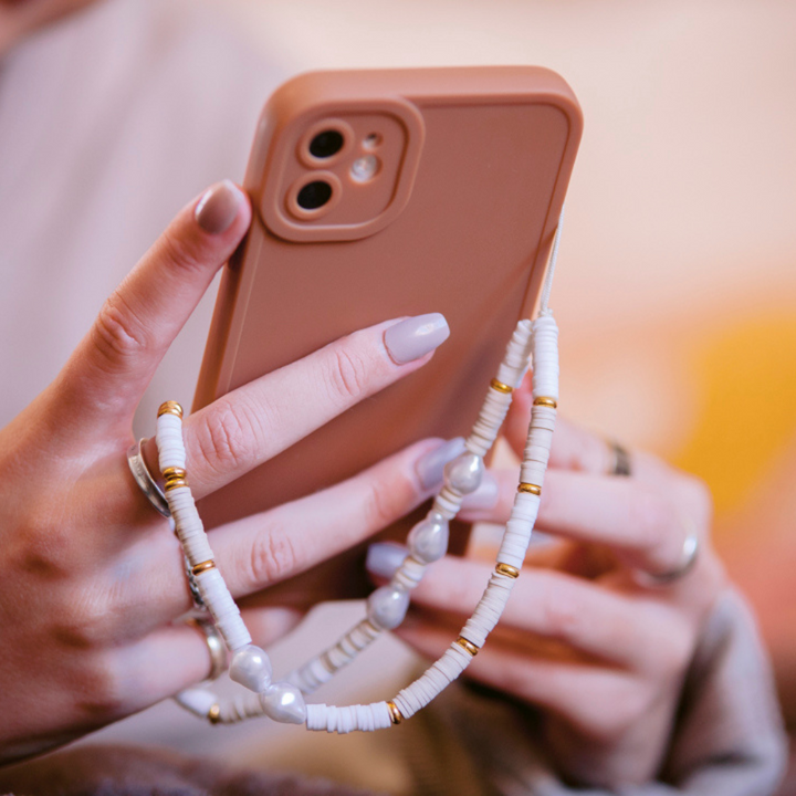 A person with pastel-colored nails holds a NAKD Phone Case - Chocolate Brown from Coconut Lane, adorned with a beaded strap. The strap, made of white and gold beads, drapes elegantly around their fingers.