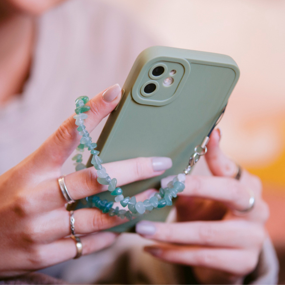 A person with neatly manicured nails, wearing multiple rings, holds a green smartphone adorned with a decorative Crystal Phone Strap - Green by Coconut Lane. The focus is on the hands and the phone, which is the center of the image.