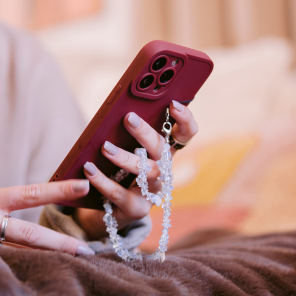 Hand with painted nails holds a maroon phone featuring a clear crystal Moonstone strap, resting on a blanket indoors.