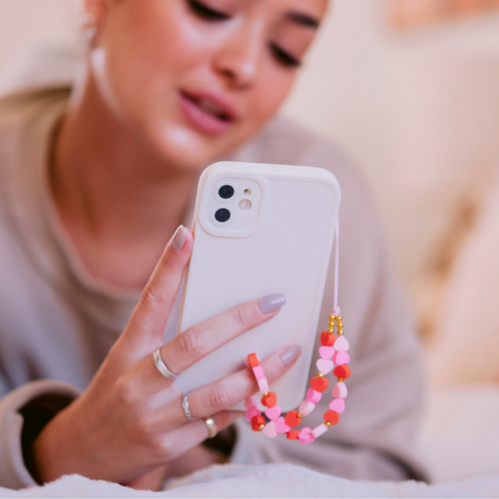 A person holds a white smartphone with a pink and orange beaded charm attached, encased in the NAKD Phone Case - Creamy White by Coconut Lane. The person is wearing a beige top, and their fingers are adorned with rings. Their face is partially visible and blurred, giving the image a soft focus.