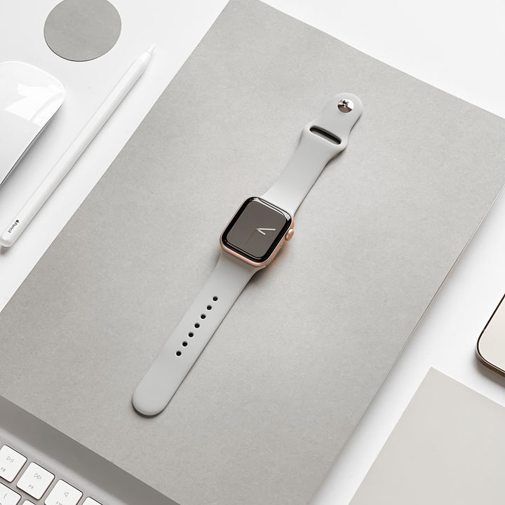 A smartwatch with a rectangular face and a NAKD Apple Watch Strap - Ice Grey by Coconut Lane is centered on a sheet of gray paper. Surrounding items include a white pen, a desktop keyboard, and part of a smartphone. The scene is minimalist and organized with a neutral color palette.