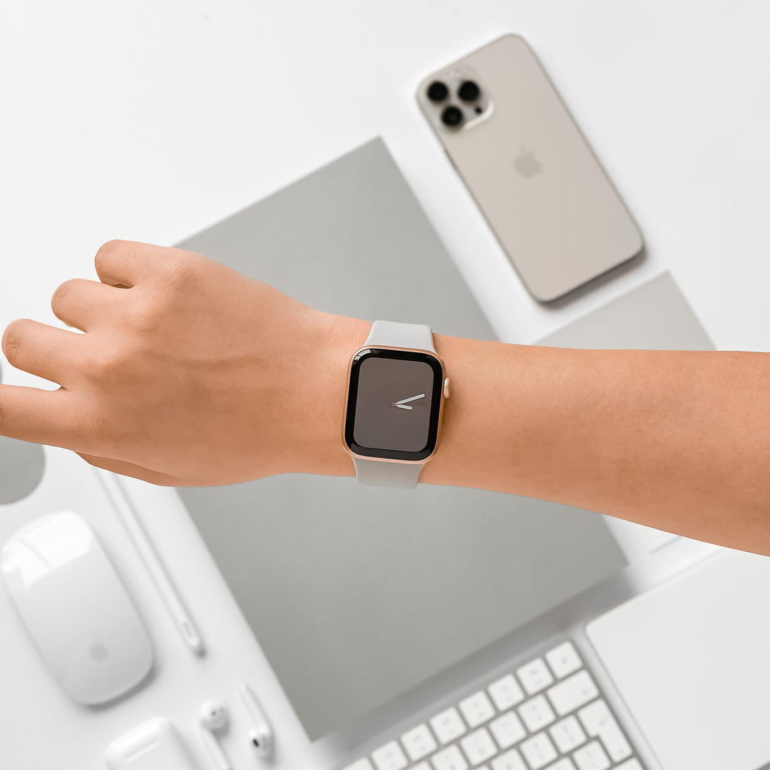 A hand wearing the NAKD Apple Watch Strap in Ice Grey by Coconut Lane, displaying a simple clock face. On the white desk are a silver smartphone, a closed gray laptop, a wireless keyboard, a mouse, earphones, a stylus, and a charger. The layout is neat and minimalistic.