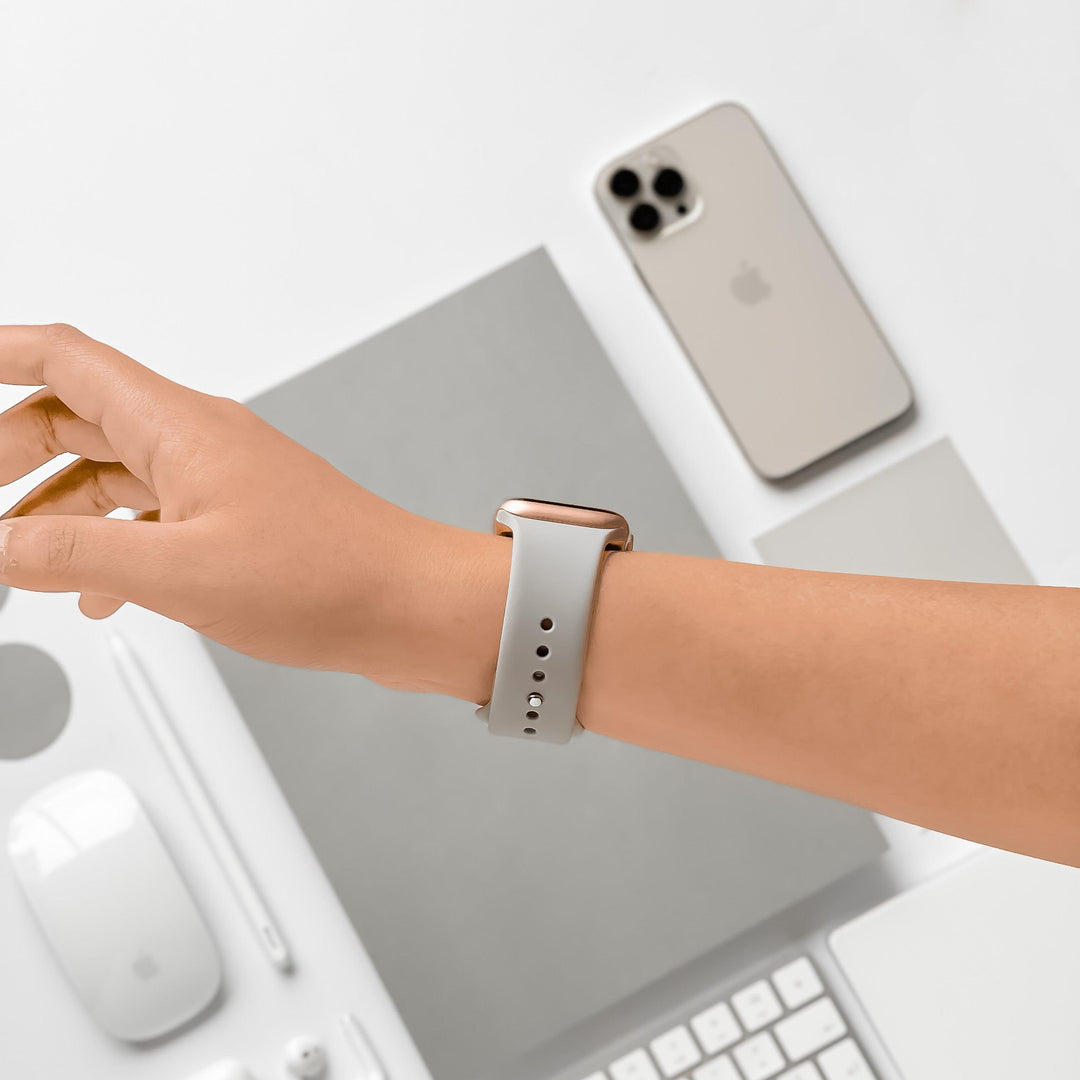 A person's hand with a Coconut Lane NAKD Apple Watch Strap in Ice Grey is extended over a white desk. The desk displays a smartphone, a keyboard, a mouse, a trackpad, a notebook, and a pen, all in white or grey tones. The setting creates a clean, minimalist workspace aesthetic.