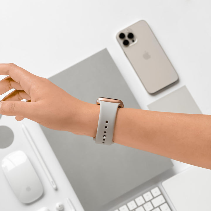 A person's hand with a Coconut Lane NAKD Apple Watch Strap in Ice Grey is extended over a white desk. The desk displays a smartphone, a keyboard, a mouse, a trackpad, a notebook, and a pen, all in white or grey tones. The setting creates a clean, minimalist workspace aesthetic.