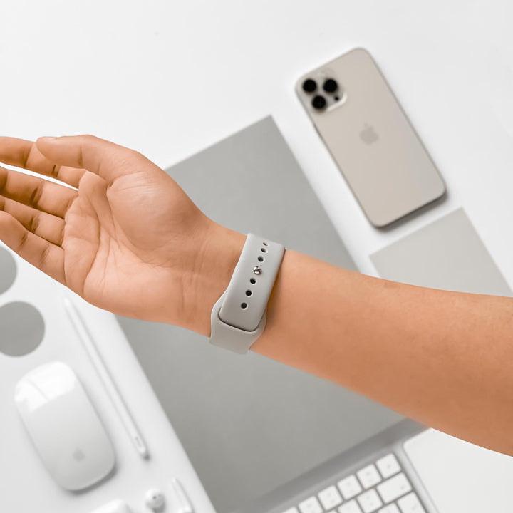 A person's wrist showcases a NAKD Apple Watch Strap - Ice Grey from Coconut Lane. The background includes a smartphone, a closed laptop, a wireless mouse, a keyboard, a stylus, and a round gray coaster, all arranged neatly on a white surface.