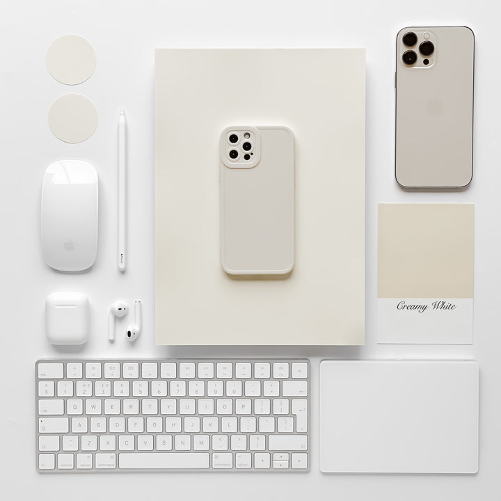 A flat lay image showcasing a collection of white tech gadgets and accessories neatly arranged on a white background. It features an iPhone with the NAKD Phone Case - Creamy White by Coconut Lane, a wireless mouse, keyboard, AirPods, Apple Pencil, round stickers, a mouse pad, and a paint swatch labeled "Creamy White.