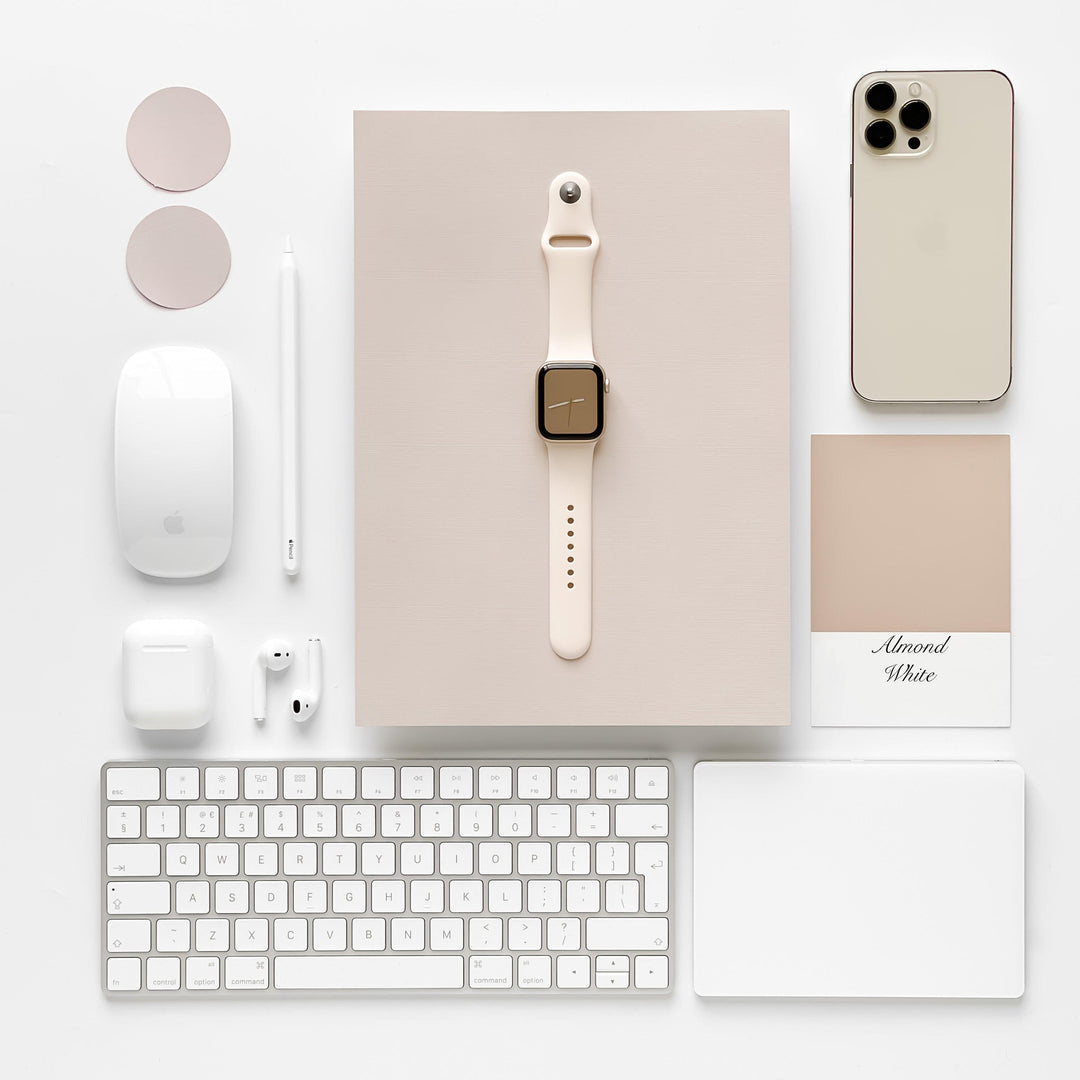 A neatly arranged flat lay features a variety of tech gadgets: an iPhone, a keyboard, a mouse, a trackpad, wireless earphones, an Apple Pencil, color swatches labeled "Almond White," and an Apple Watch with the sleek NAKD Apple Watch Strap in Almond White by Coconut Lane—all set against a beige-colored background.