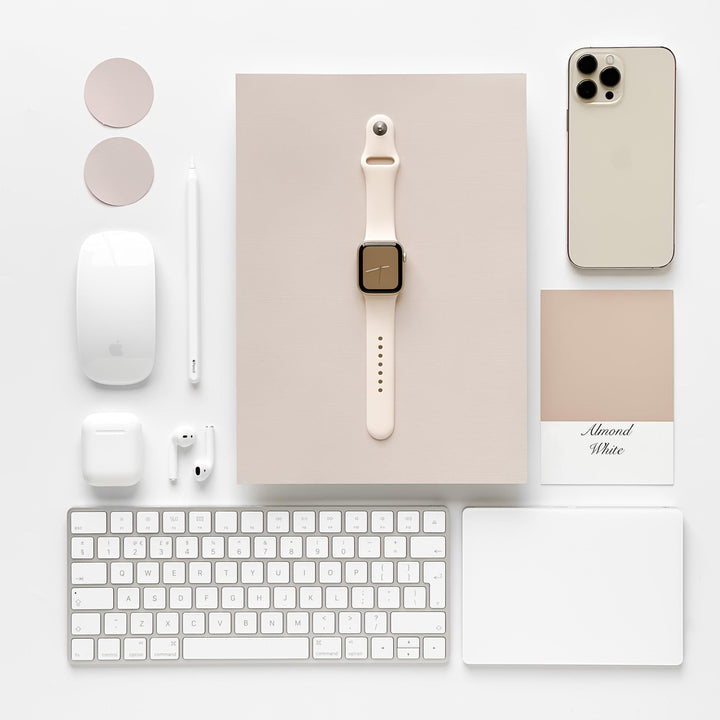 A neatly arranged flat lay features a variety of tech gadgets: an iPhone, a keyboard, a mouse, a trackpad, wireless earphones, an Apple Pencil, color swatches labeled "Almond White," and an Apple Watch with the sleek NAKD Apple Watch Strap in Almond White by Coconut Lane—all set against a beige-colored background.