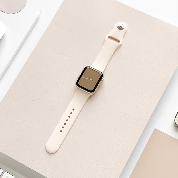 The NAKD Apple Watch Strap - Almond White by Coconut Lane is displayed on a light beige surface. The background features a white stylus, a small portion of a light-colored laptop, and other beige and white items, creating a minimalistic and elegant aesthetic.