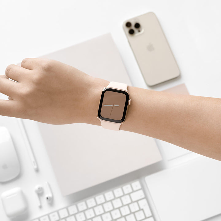 A person's hand is shown wearing an Apple Watch with the NAKD Apple Watch Strap in Almond White by Coconut Lane. The background features a beige smartphone with three cameras, a keyboard, a pair of earbuds, and a mouse, all in a minimalist and neutral-toned workspace setup.