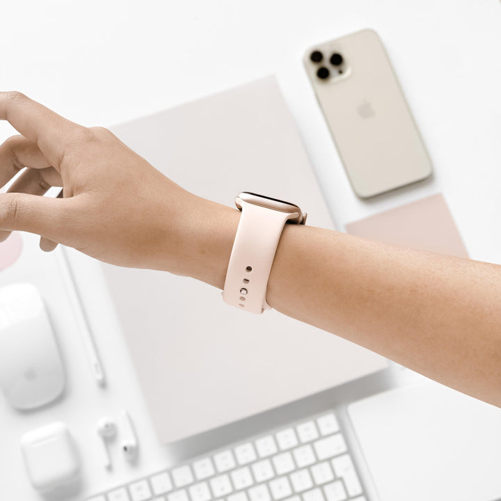 A person's hand, wearing an Almond White NAKD Apple Watch Strap by Coconut Lane, is extended slightly above a desk. The desk holds a beige smartphone, an Apple Watch model, closed laptop, wireless earbuds, keyboard, mouse, and stylus—all in a minimalist, neutral-toned color scheme.