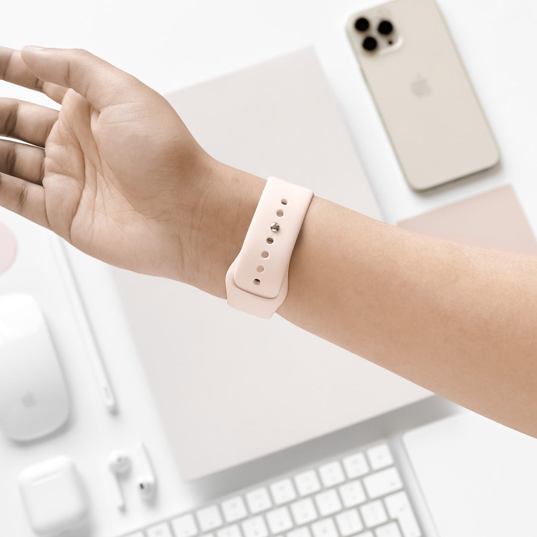 A person wearing an Apple Watch with the NAKD Apple Watch Strap in Almond White by Coconut Lane on their wrist. In the background, there is a gold smartphone, white keyboard, computer mouse, earbuds and a notepad, all arranged on a white surface. The setting is minimalistic and clean.