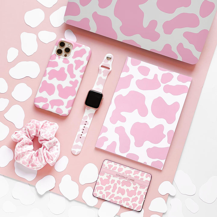 A flat lay of various tech and accessory items with a pink cow print pattern. Items include a MacBook, iPhone case, notebook, scrunchie, and a rectangular card. The Pink Cow Apple Watch Strap by Coconut Lane perfectly complements the coordinated background with the same pink and white cow print.