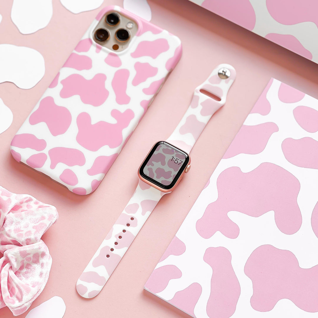 A flat lay image showcasing a smartphone with a pink and white cow-print case, the Coconut Lane Pink Cow Apple Watch Strap in matching silicone, a scrunchie, and various cow-print patterned items in pink and white, all neatly arranged on a pink surface.