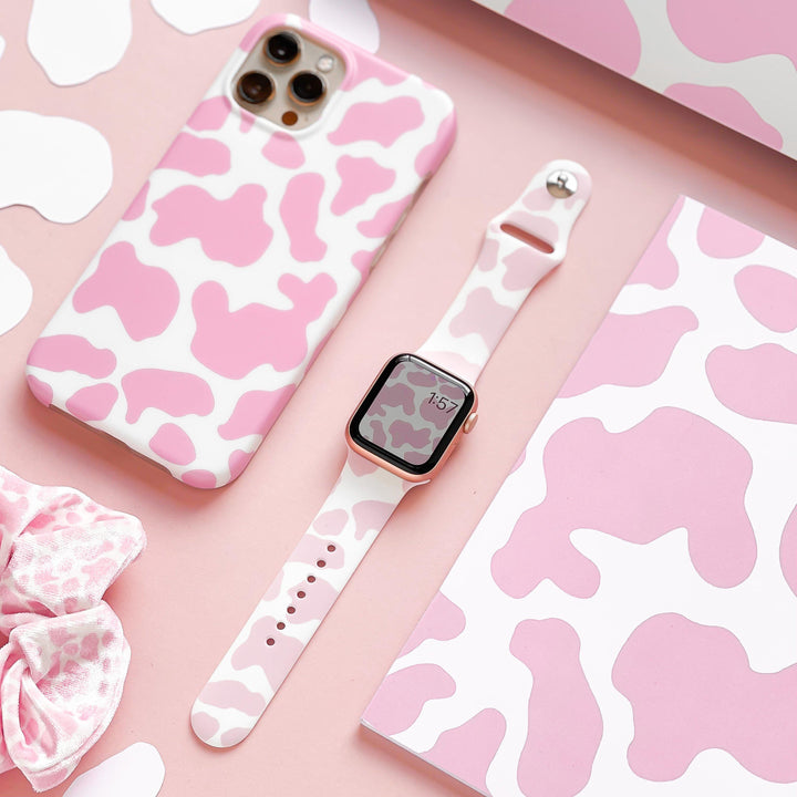 A flat lay image showcasing a smartphone with a pink and white cow-print case, the Coconut Lane Pink Cow Apple Watch Strap in matching silicone, a scrunchie, and various cow-print patterned items in pink and white, all neatly arranged on a pink surface.