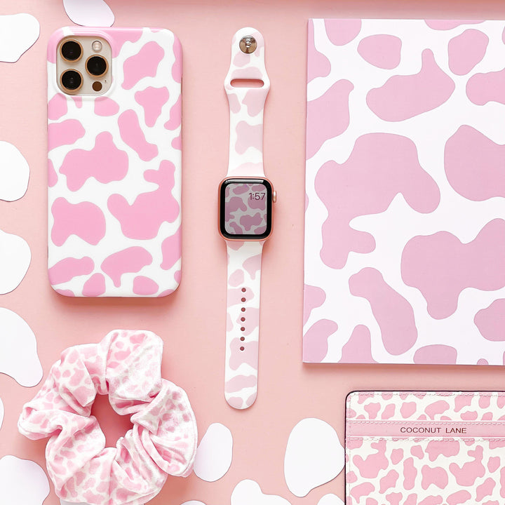 A flat lay of Coconut Lane accessories featuring a pink and white cow print design, includes an iPhone case, the Pink Cow Apple Watch Strap, a scrunchie, a notebook, and a wallet. The background is a matching pink color that enhances the cohesive aesthetic of the items.
