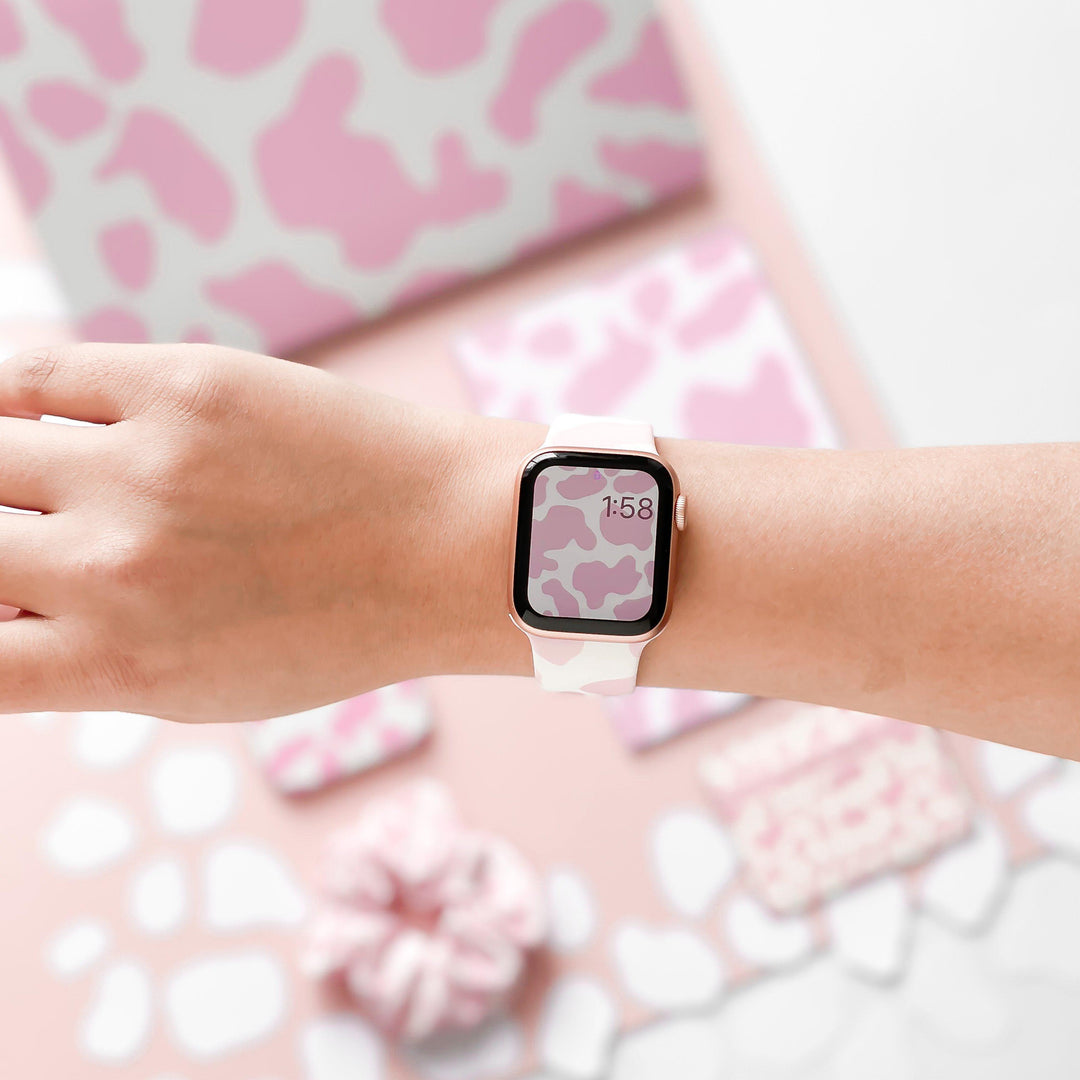 A person's wrist wearing an Apple Watch with Coconut Lane's Pink Cow Apple Watch Strap and a camo watch face. In the background, matching pink and white camo objects, including a notebook, phone case, and scrunchie, are visible. The time on the watch reads 1:58.
