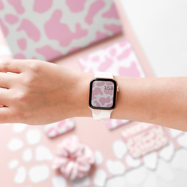 A person's wrist wearing an Apple Watch with Coconut Lane's Pink Cow Apple Watch Strap and a camo watch face. In the background, matching pink and white camo objects, including a notebook, phone case, and scrunchie, are visible. The time on the watch reads 1:58.