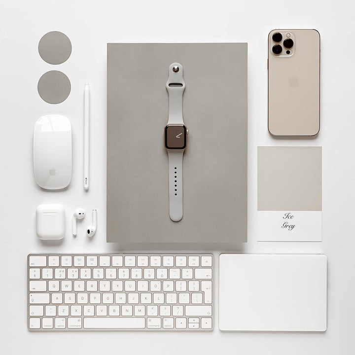 A neatly arranged collection of tech gadgets and accessories on a white surface, featuring an iPhone, Apple Watch with a NAKD Apple Watch Strap - Ice Grey by Coconut Lane, AirPods, a Mac keyboard, Magic Mouse, Apple Pencil, and trackpad. A paint sample card labeled "Ice Grey" and two round grey stickers are also included.