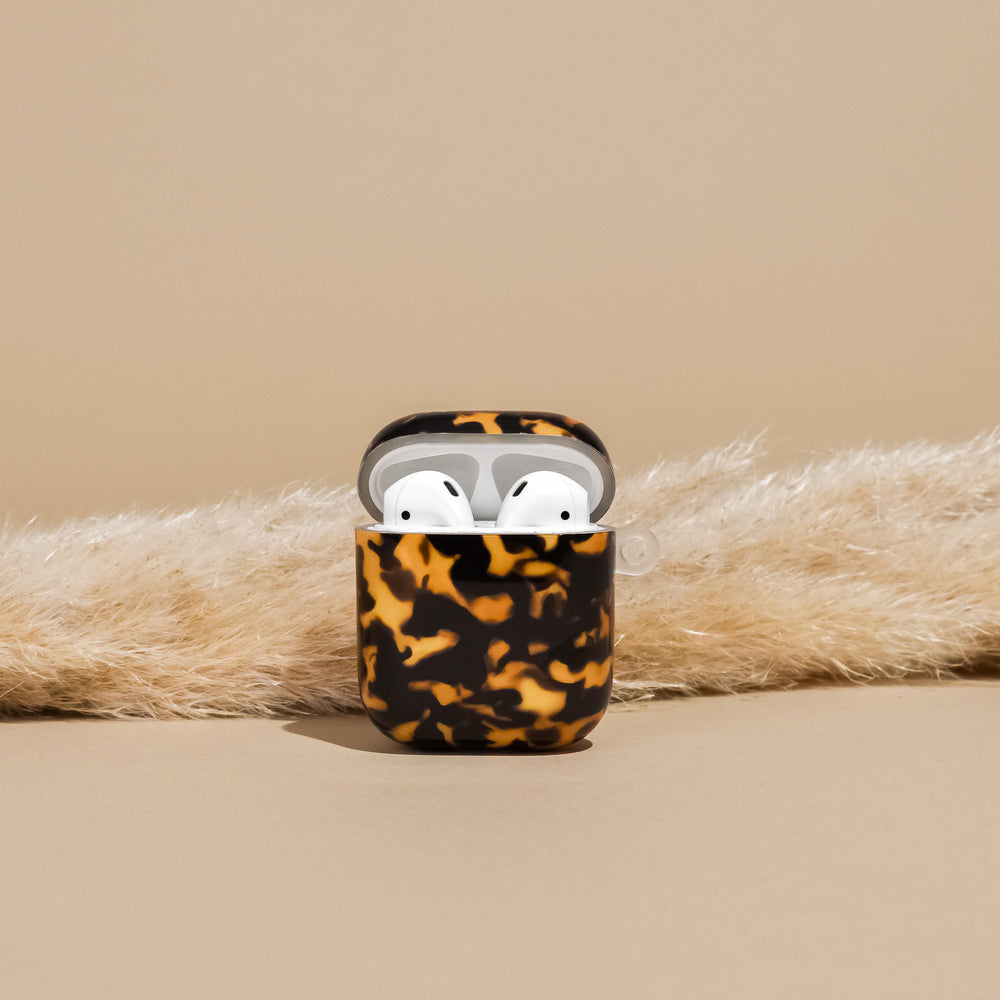 Tortoiseshell AirPods case by Coconut Lane on a beige surface with pampas grass in the background.