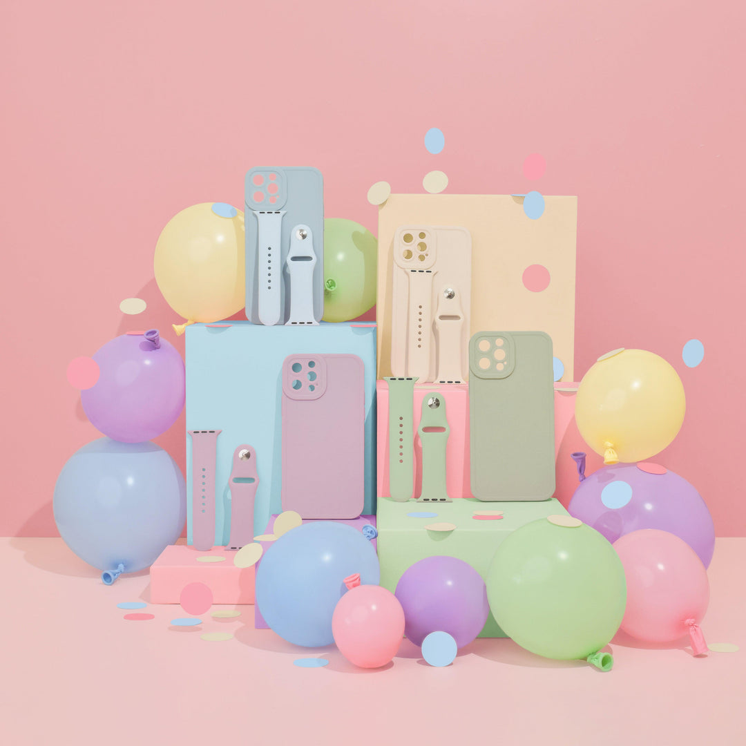 A display of vibrant iPhone cases and NAKD Apple Watch Straps in Almond White by Coconut Lane, featuring silicone straps arranged on pastel-colored boxes, surrounded by balloons in various sizes and colors. The backdrop is pink, and confetti is scattered around the arrangement, creating a festive atmosphere perfect for showcasing the NAKD Drop 2 collection.