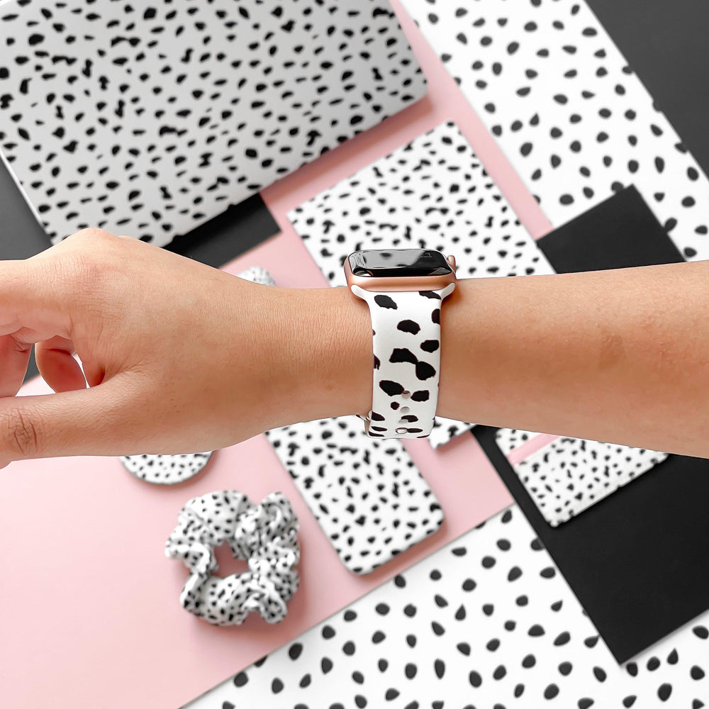 A hand is shown wearing the Coconut Lane Monochrome Spots Apple Watch Strap, showcasing a white band adorned with black irregular spots. The background features an assortment of items, including a scrunchie, phone cases, and canvases, all displaying the same spotted pattern from the Monochrome Spots Collection, arranged on a pink and black surface.
