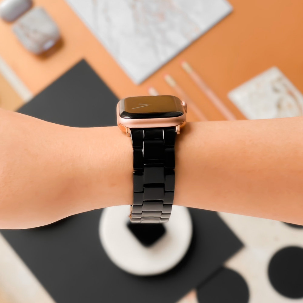 Close-up view of an arm wearing Coconut Lane's Luxe Black Apple Watch strap paired with a rose gold casing. The background showcases a minimalistic desk setup, featuring a laptop, a marble notebook, and stationery, all contributing to an overall warm and tidy aesthetic.
