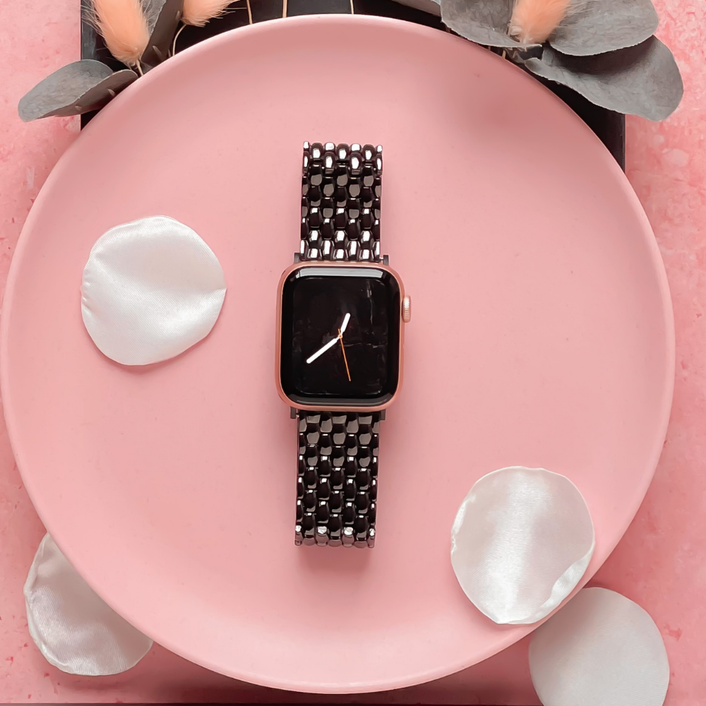 A smartwatch fitted with a Coconut Lane Mermaid Apple Watch Strap - Black and a metal link band is placed on a pink plate. The watch face shows the time. White, silk-like petals beautify the plate, which rests on a textured pink background. Gray and blush decorative foliage encircle the arrangement.