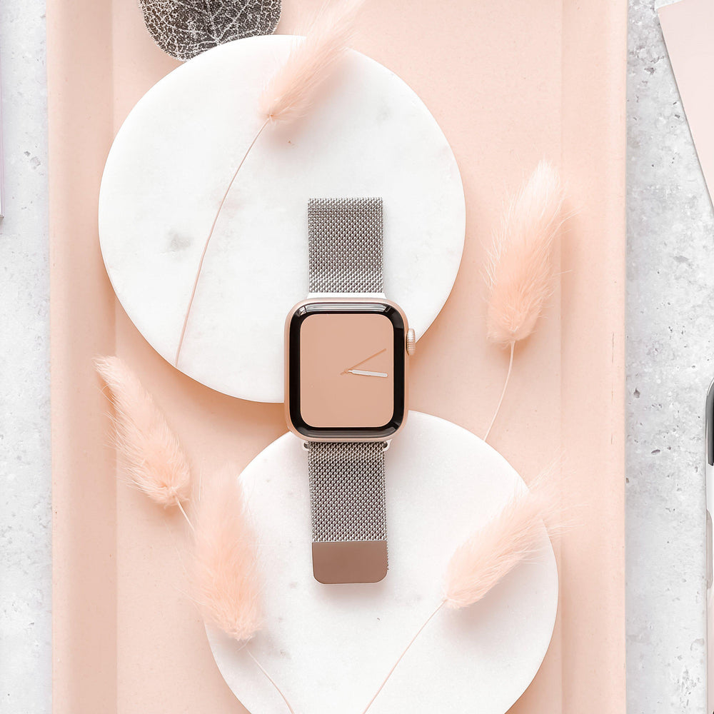 The Coconut Lane Magnetic Chain Apple Watch Strap in Silver rests stylishly across two round marble trays. The background showcases delicate, peach-colored feathery plants, creating a soft and elegant aesthetic. The watch face displays a minimalistic peach-colored design, compatible with various Apple Watch models.